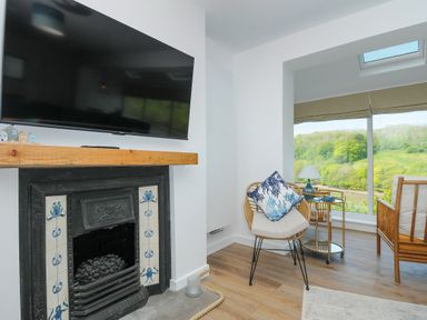 A sitting area with a TV above a fireplace a chair with a cushion a side table with a lamp and a large window showing greenery at Anjoma in Looe