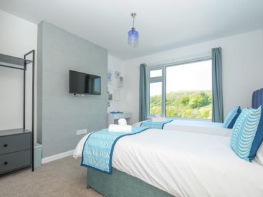 A bedroom with two single beds blue patterned pillows and throws a wall mounted TV and a large window overlooking trees at Anjoma in Looe