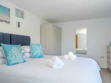 A bedroom with a bed with blue headboard and turquoise sea-themed pillows a wardrobe a mirror and a chest of drawers at Anjoma in Looe