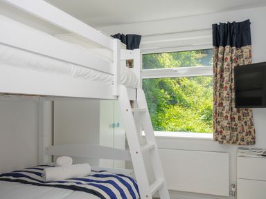 A bedroom with a white bunk bed a window with patterned curtains and a wall mounted television at Anjoma in Looe