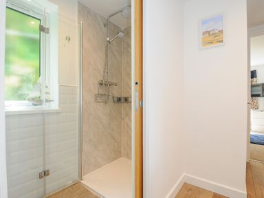 A shower area with glass door and window beside a hallway with wooden floor and framed picture on the wall at Anjoma in Looe
