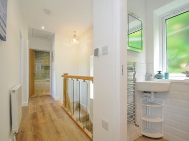 A hallway with wooden floor leading to a bathroom and stairs with a glass and wood railing at Anjoma in Looe