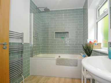 A bathroom with a bathtub and shower with green tiles on the walls and a window with plants at Anjoma in Looe