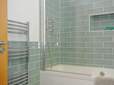 A bathroom with green tiled walls a bathtub a handheld shower a glass shower screen and a chrome towel radiator at Anjoma in Looe