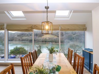 A dining room with a wooden table and chairs beside large windows overlooking a river view at Anjoma in Looe