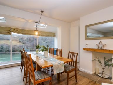 A dining room with wooden table and chairs by large windows overlooking a river at Anjoma in Looe