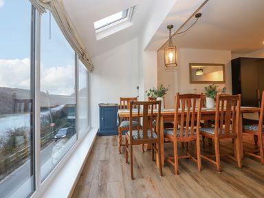 A dining area with a wooden table and chairs next to large windows overlooking a river at Anjoma in Looe