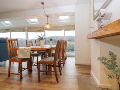 A dining room with a wooden table and chairs near large windows with beige blinds at Anjoma in Looe
