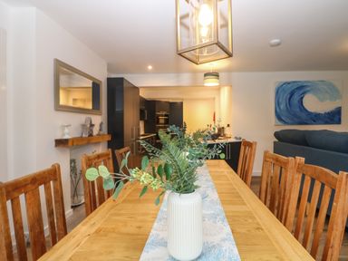 A dining area with wooden table and chairs a vase with green foliage on the table a kitchen with black cabinets in the background and a blue wave painting on the wall at Anjoma in Looe