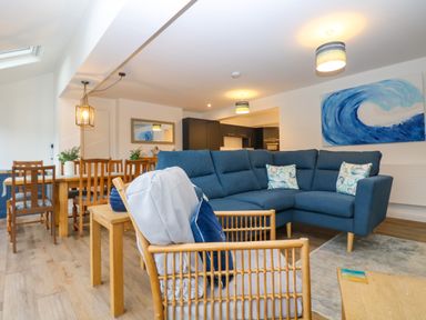 A living room with blue sofa wooden dining table and chairs with wave painting on the wall at Anjoma in Looe