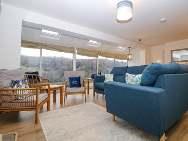 A living room with blue sofas wooden chairs and large windows overlooking trees at Anjoma in Looe