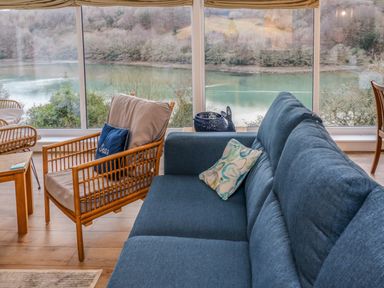 A living room with a blue sofa and a wooden armchair with cushions by large windows overlooking a river and trees at Anjoma in Looe