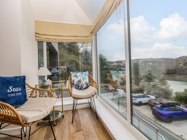 A sunroom with wicker chairs cushions lamp and a small table by large windows overlooking a parking lot and water at Anjoma in Looe