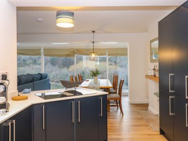 A kitchen with dark cabinets and a dining area with wooden chairs and a table near large windows at Anjoma in Looe