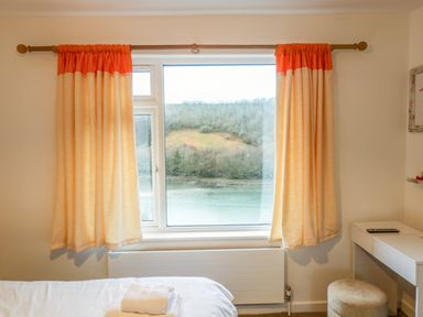A bedroom window with orange and cream curtains overlooking a river and hills at Anjoma in Looe