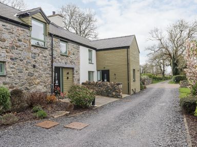 Stone and wood exterior houses beside a gravel driveway with plants and trees at Gwalia Cottage in Brynsiencyn