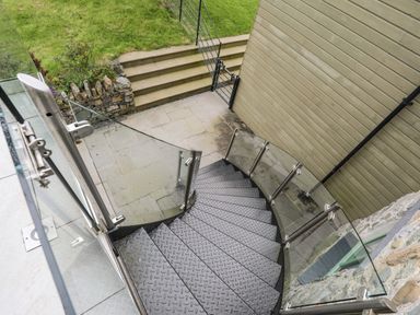 A metal spiral staircase with glass railings leading down to a stone patio with steps and grass at Gwalia Cottage in Brynsiencyn