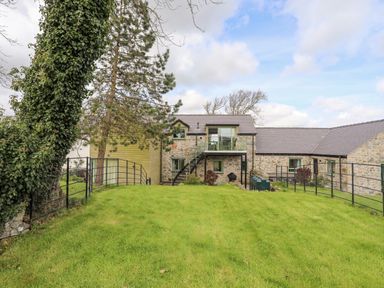 A stone house with a balcony and staircase in a grassy fenced yard at Gwalia Cottage in Brynsiencyn