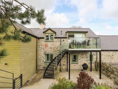 A stone and wooden house with an outdoor staircase leading to a glass balcony with a table and chairs at Gwalia Cottage in Brynsiencyn
