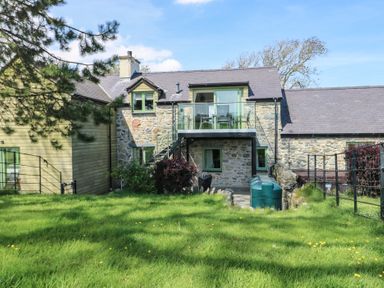 A stone and wood exterior of a two story house with a glass balcony and green grass lawn at Gwalia Cottage in Brynsiencyn