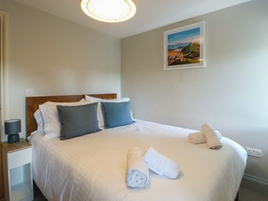 A bedroom with a double bed with white bedding and gray pillows a bedside table with a lamp and a framed landscape picture on the wall at Gwalia Cottage in Brynsiencyn