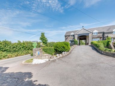 A stone building with an archway and a driveway lined with hedges and bushes at The Hayloft in St Florence