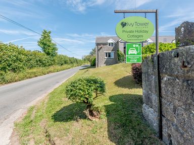 A road next to a grassy area with a stone wall and sign for Ivy Tower Village with cottages and electric vehicle charging at The Hayloft in St Florence