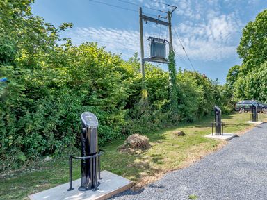 Outdoor electric vehicle charging points near bushes and trees with a power transformer overhead and a parked car at The Hayloft in St Florence