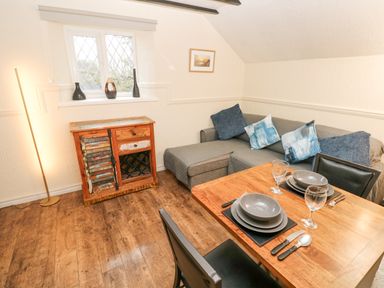 A dining area with a wooden table set for two next to a gray sofa with cushions and a wooden cabinet under a window at The Hayloft in St Florence