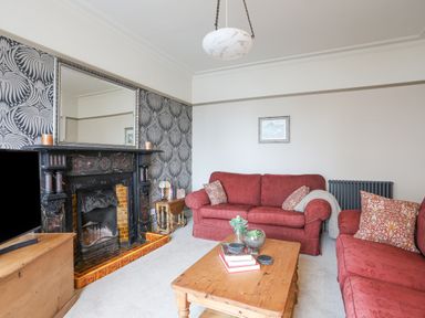A living room with red sofas a wooden coffee table a fireplace with a large mirror above and patterned wallpaper at Llwyn Onn in Criccieth