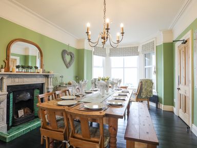 A dining room with a wooden table set with plates and glasses a fireplace and a window with floral curtains at Llwyn Onn in Criccieth