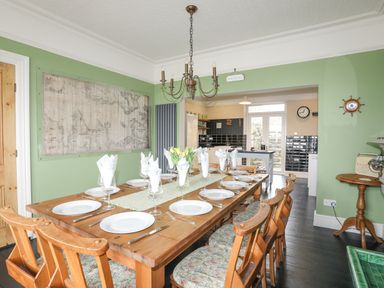 A dining room with a set table and wooden chairs next to a kitchen at Llwyn Onn in Criccieth