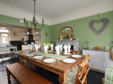 A dining room with a wooden table set with plates and glasses beside a green wall with a heart decoration at Llwyn Onn in Criccieth