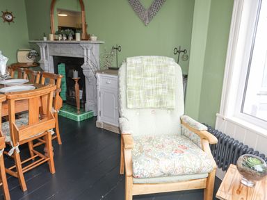 A dining area with wooden chairs and a table beside a living room with an armchair a fireplace and a window at Llwyn Onn Criccieth
