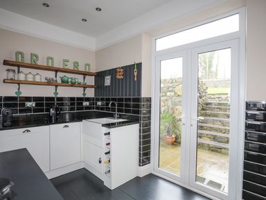 A kitchen with black countertops white cabinets open wine storage and glass doors leading outside at Llwyn Onn in Criccieth