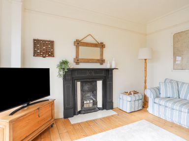 A living room with a wooden floor a black fireplace a TV on a wooden stand a striped sofa and ottoman a floor lamp and wall mirror at Llwyn Onn in Criccieth