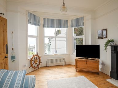 A living room with wooden floors a bay window striped blinds a television on a wooden cabinet and a ship wheel at Llwyn Onn in Criccieth
