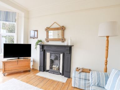 A living room with a black fireplace, wooden floor, TV on a wooden chest, blue striped sofa, and a tall floor lamp at Llwyn Onn in Criccieth