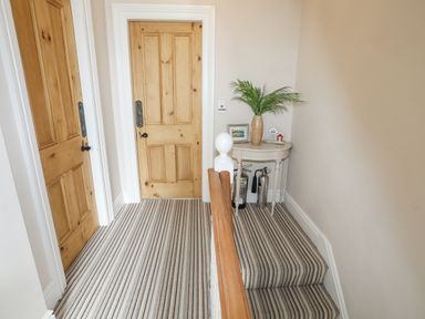 A hallway with two wooden doors a small table with a plant framed picture and decorative items striped carpet on stairs and floor at Llwyn Onn in Criccieth