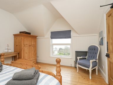 A bedroom with a wooden bed with towels on it a wooden wardrobe a small wooden table with a model boat and a blue cushioned armchair at Llwyn Onn in Criccieth