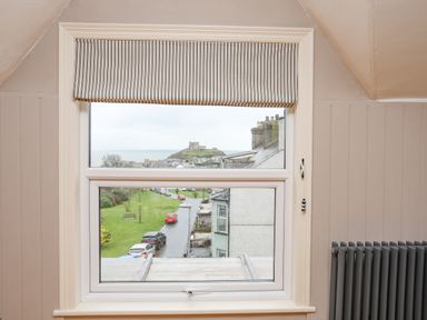 A window with a striped blind partially open showing a street and a castle on a hill at Llwyn Onn in Criccieth