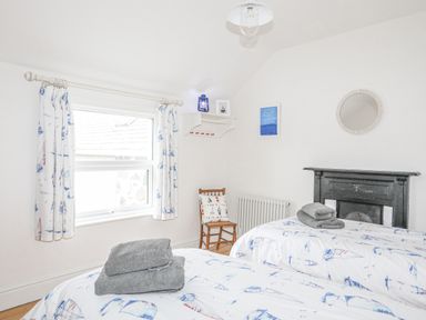 A bedroom with twin beds and gray towels on the beds a wooden chair by the window a black fireplace a round mirror on the wall and curtains with sailboat patterns at Llwyn Onn in Criccieth