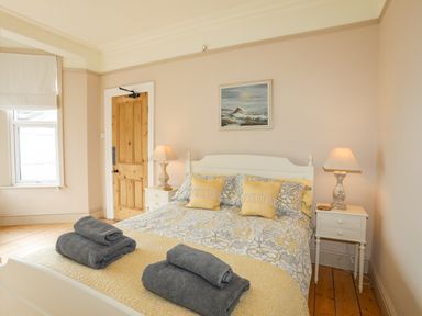 A bedroom with a bed with patterned bedding and pillows two nightstands with lamps wooden door and window at Llwyn Onn in Criccieth