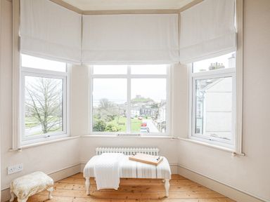A bay window with white blinds two stools and a radiator with a view of a street and castle at Llwyn Onn in Criccieth