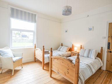 A bedroom with two single wooden beds, a wicker chair with cushion, a window with striped blind and wooden floor at Llwyn Onn in Criccieth