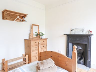 A bedroom with a wooden bed, chest of drawers with a mirror, clothes hanger rack, and a black fireplace at Llwyn Onn in Criccieth