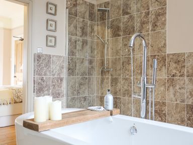 A bathroom with a white bathtub with wood tray holding candles and lotion bottle next to a tiled wall with shower at Llwyn Onn in Criccieth
