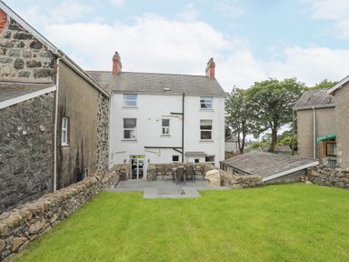 A backyard with a lawn and stone patio with chairs and a table behind a white house at Llwyn Onn in Criccieth