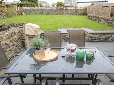 A patio with a glass table set with a teapot cups a tray with glasses and a plant beside a grassy garden at Llwyn Onn in Criccieth