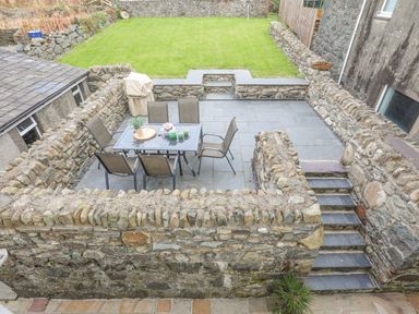 An outdoor stone patio with six chairs around a glass table and a covered grill next to a grassy lawn at Llwyn Onn in Criccieth